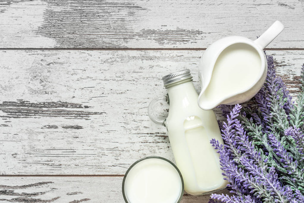 Vintage glass bottle with milk, a glass with milk and a beautiful white jug next to a branch of lavender flowers on a vintage wooden background. View from above. - Valokuva, kuva