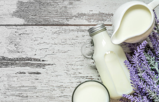Vintage glass bottle with milk, a glass with milk and a beautiful white jug next to a branch of lavender flowers on a vintage wooden background. View from above. - Foto, Imagem