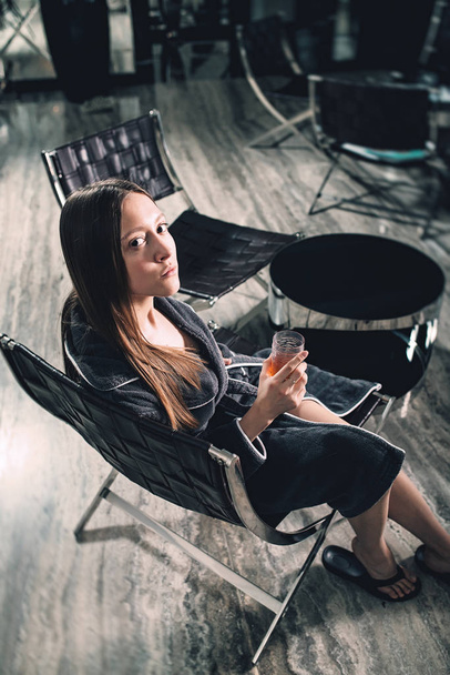 Portrait of young beautiful woman relaxing in a chair in a bathrobe with a detox drink in a hand. Luxery spa center. look at the camera - Foto, Imagen