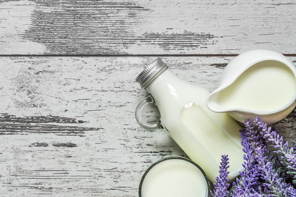 Vintage glass bottle with milk, a glass with milk and a beautiful white jug next to a branch of lavender flowers on a vintage wooden background. View from above. - Photo, image