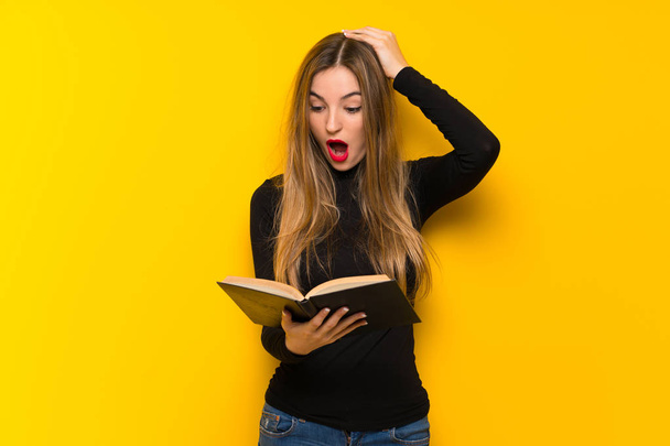 Young pretty woman over yellow background surprised while enjoying reading a book - Photo, image