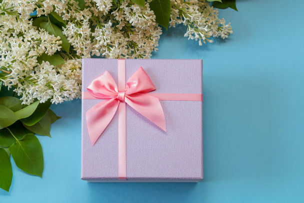 Geschenkbox mit rosa Schleife und Blumen aus weißem Flieder auf blauem Hintergrund. Ansicht von oben, Kopierraum. - Foto, Bild
