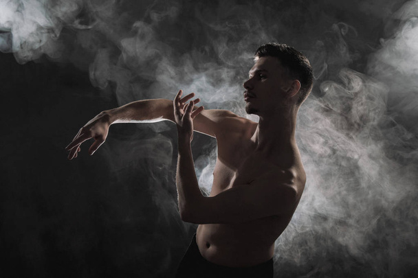 A young male ballet dancer with black leggings and a naked torso performs dance moves against a gray grunge background, with a light of lights and smoke. - Foto, Imagem