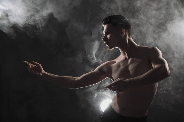A young male ballet dancer with black leggings and a naked torso performs dance moves against a gray grunge background, with a light of lights and smoke. - Valokuva, kuva