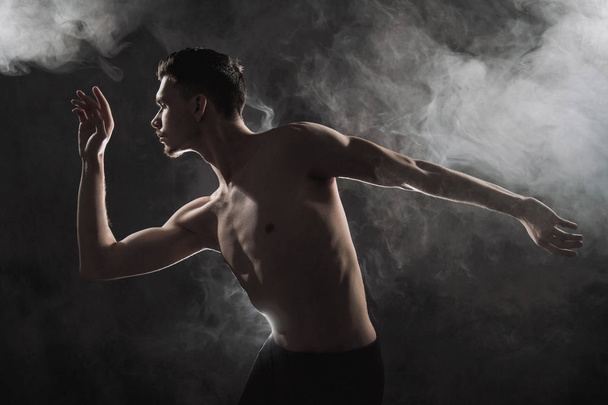 A young male ballet dancer with black leggings and a naked torso performs dance moves against a gray grunge background, with a light of lights and smoke. - Photo, Image
