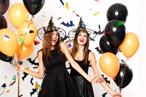 Two beautiful girls in black dresses and witch hats have fun with black and orange balloons and confetti.Halloween party . - Φωτογραφία, εικόνα