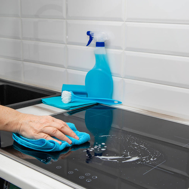 A woman's hand with a blue microfiber cloth rubs a glass ceramic plate in the kitchen. In the background-are cleaning products. - Photo, image