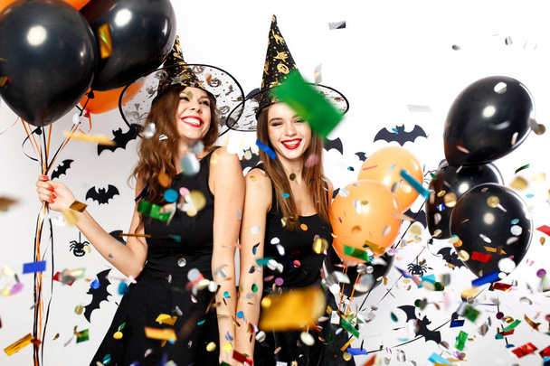Two beautiful girls in black dresses and witch hats have fun with black and orange balloons and confetti. Halloween party - Foto, Imagen