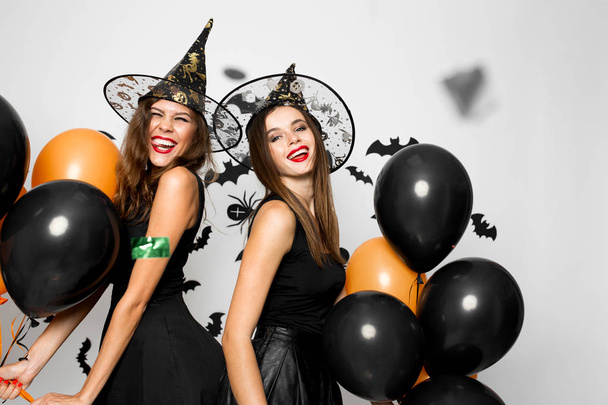 Two beautiful girls in black dresses and witch hats have fun with black and orange balloons. Halloween - 写真・画像