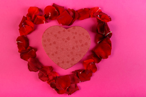 Box with a gift in the shape of a heart surrounded by rose petals on a pink background. - Fotó, kép