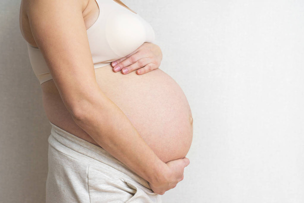 Schwangere hält Hände auf Bauch vor hellem Hintergrund. Schwangerschaft, Mutterschaft, Vorbereitung und Erwartung Konzept. Nahaufnahme, Kopierraum, drinnen. schöne zarte Stimmung Foto der Schwangerschaft - Foto, Bild