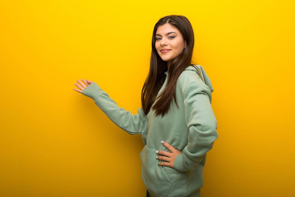 Teenager girl with green sweatshirt on yellow background pointing back and presenting a product - Foto, immagini