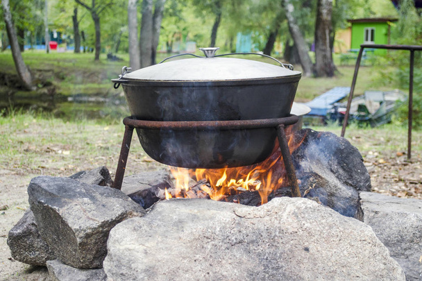 Large metal pot with a lid and handle on tripod used for cooking. Cooking in a bowler hat on the fire in nature - Photo, Image