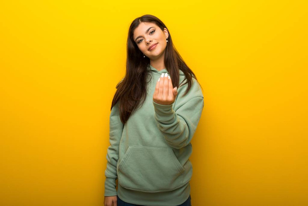 Teenager girl with green sweatshirt on yellow background inviting to come with hand. Happy that you came - Foto, afbeelding