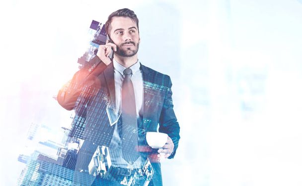 Handsome bearded young businessman talking on smartphone holding cup of coffee over blue cityscape background. Toned image double exposure mock up - Photo, image
