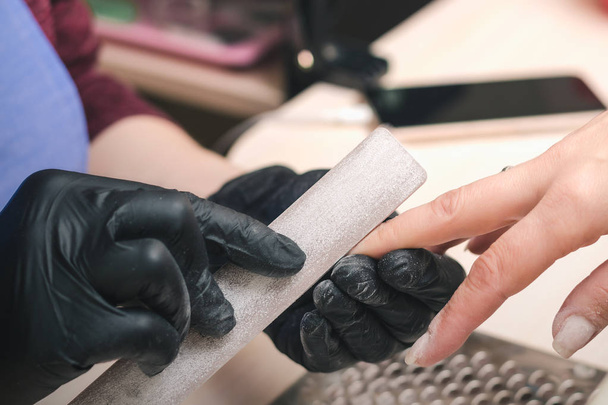 Manicurist in black latex gloves form the shape of a client's nail in a beauty salon. - 写真・画像