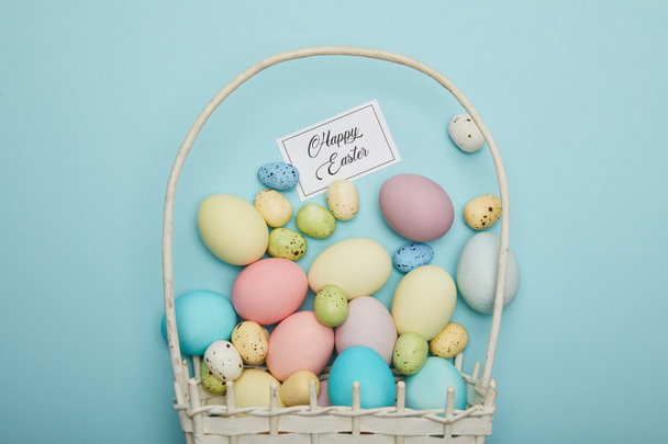 Top view of painted easter chicken and quail eggs in straw basket, card with happy easter lettering - Fotó, kép