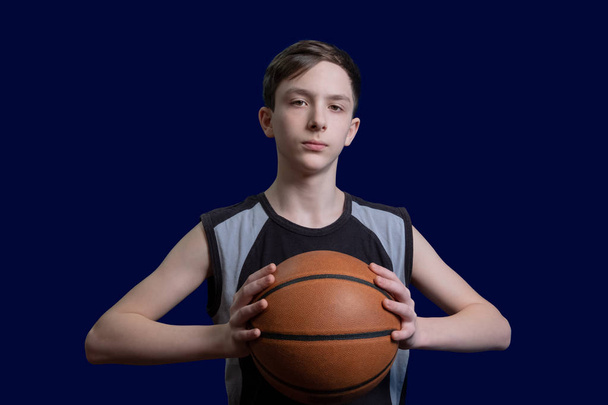 Portrait of a teen basketball player. The guy in the black t-shirt holds the ball in his hands. The concept of a sports poster or design news about basketball competitions. Isolated on blue background. - Fotoğraf, Görsel