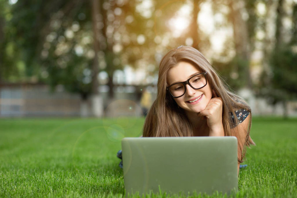 Happy teenage girl in the glasses looking at the notebook outdoors lying down on green lawn headphones on head. Multicultural model, mixed race, asian russian girl. Copy space. People and technology. - Zdjęcie, obraz