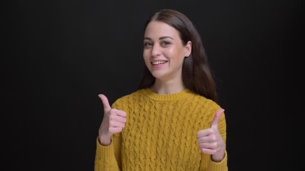 Retrato de chica morena de pelo largo en suéter amarillo haciendo un gesto de dos signos de dedo hacia arriba para mostrar como y respeto sobre fondo negro
. - Metraje, vídeo