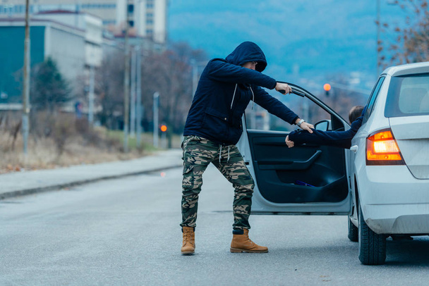 The car thief is pulling the car owner out of his car and trying to get the car while pointing a loaded gun at the drivers head. - Photo, Image