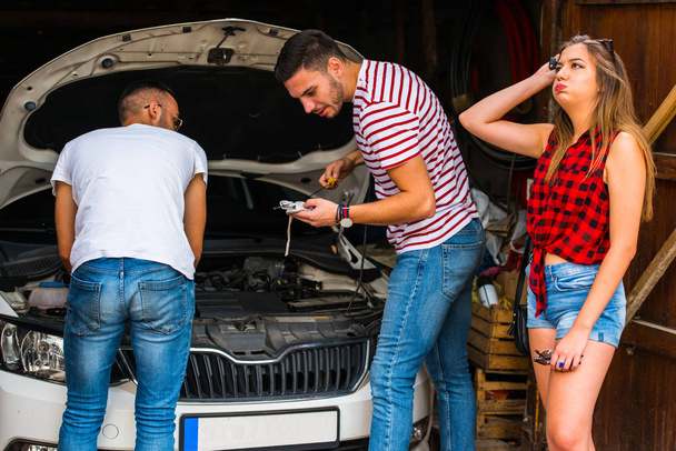 Dois caras tentando consertar um troble de carro enquanto uma garota está ao lado deles
 - Foto, Imagem