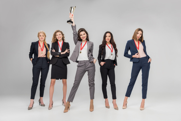 cheerful businesswoman holding trophy above head and standing near attractive colleagues posing on grey background - Photo, Image