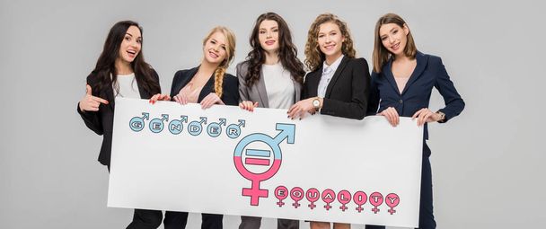 cheerful young woman pointing with finger at large sign with gender equality symbol near female corowokers isolated on grey  - Photo, Image