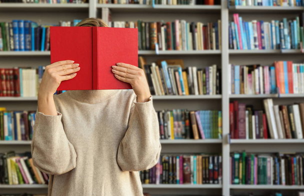 Mujer joven escondiendo la cara detrás del libro en la biblioteca moderna
 - Foto, imagen