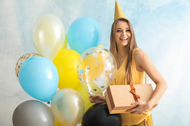 Красивая молодая женщина с воздушными шарами, шляпа вечеринки и подарок на светлом фоне
 - Фото, изображение