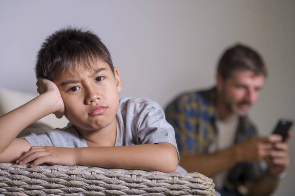 young sad and bored 7 or 8 years old child at home couch feeling frustrated and unattended while man networking on mobile phone as internet addict father neglecting his young son - 写真・画像