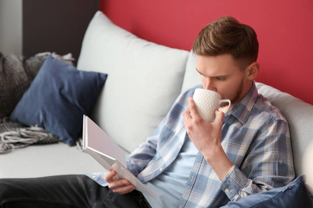 Young man drinking coffee while reading book at home - Foto, Imagem