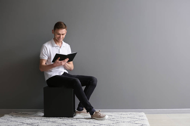 Komea nuori mies lukemassa kirjaa istuessaan lähellä harmaata seinää
 - Valokuva, kuva