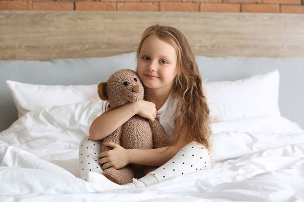 Симпатична маленька дівчинка з іграшковим ведмедем сидить на ліжку
 - Фото, зображення
