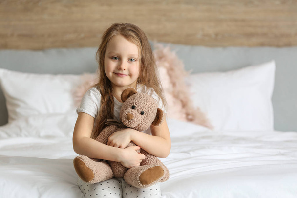 Симпатична маленька дівчинка з іграшковим ведмедем сидить на ліжку
 - Фото, зображення