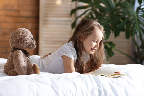 Ładna dziewczynka z zabawki opatrzone czytając książkę leżąc na łóżku - Zdjęcie, obraz