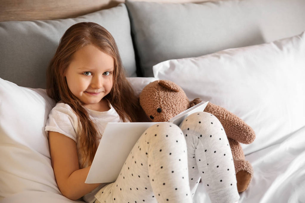 Cute little girl with toy bear reading book in bed - Photo, Image