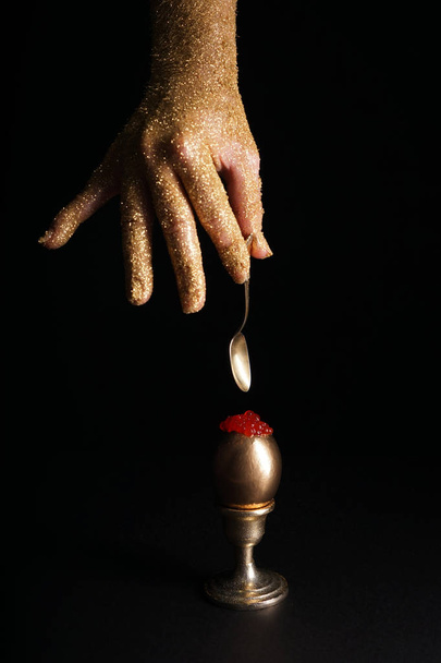 Γυναικείο χέρι κρατά κουτάλι και χρυσό αυγό με κόκκινο χαβιάρι σε σκούρο φόντο - Φωτογραφία, εικόνα