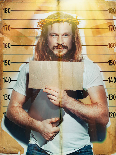 Male criminal suspect in the image of Jesus holds a laptop and a bottle of alcohol in his hand, is photographed against the background of the growth wall. Creative religious concept - Foto, afbeelding