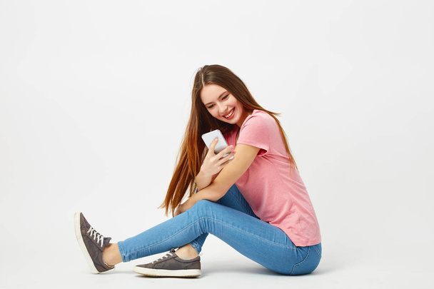Beautiful girl dressed in a pink t-shirt, jeans and sneakers sits on the floor and uses mobile phone on a white background in the studio - Φωτογραφία, εικόνα