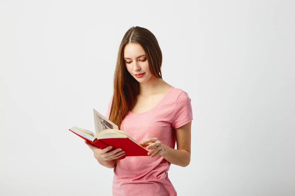 Beautiful long haired girl dressed in a pink t-shirt reads a book on a white background in the studio - Foto, imagen