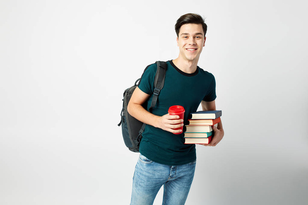 Smiling dark-haired guy with a black backpack on his shoulder dressed in a black t-shirt and jeans holds books and a red plastic cup in his hands on a white background - Φωτογραφία, εικόνα