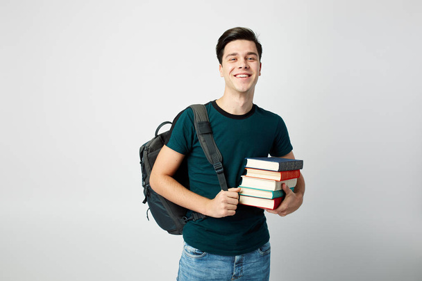 Dark-haired guy with a black backpack on his shoulder dressed in a dark t-shirt and jeans holds books in his hands on a white background - Фото, изображение