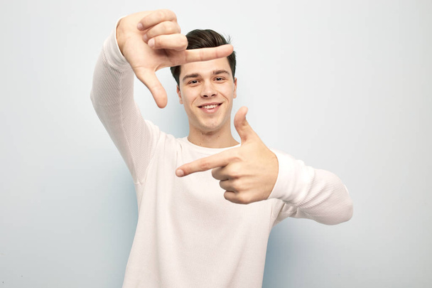 Funny dark-haired guy dressed in a white long sleeve t-shirt stands and keeps his hands in front of him making with fingers a shape of a frame on a white background - Fotografie, Obrázek