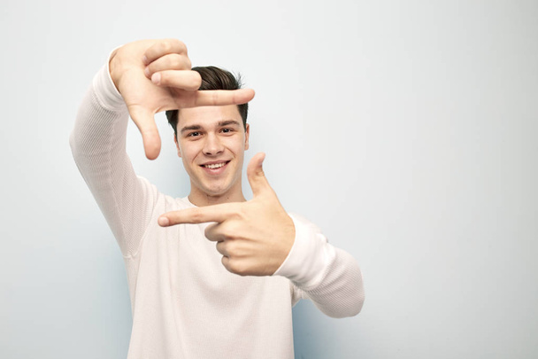 Funny dark-haired guy dressed in a white long sleeve t-shirt stands and keeps his hands in front of him making with fingers a shape of a frame on a white background - Foto, Bild