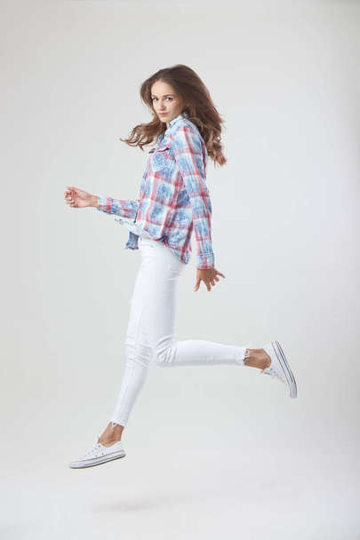 Joyful girl dressed in a checkered shirt and white jeans jumps on the white background in the studio - Fotoğraf, Görsel