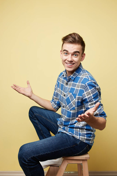 Cheerful and surprised guy dressed in a plaid shirt and jeans is sitting on a stool on a yellow background in the studio - 写真・画像