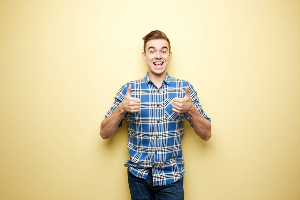 Cheerful guy dressed in a plaid shirt and jeans stands and giving thumbs up next to yellow wall in the studio - Valokuva, kuva