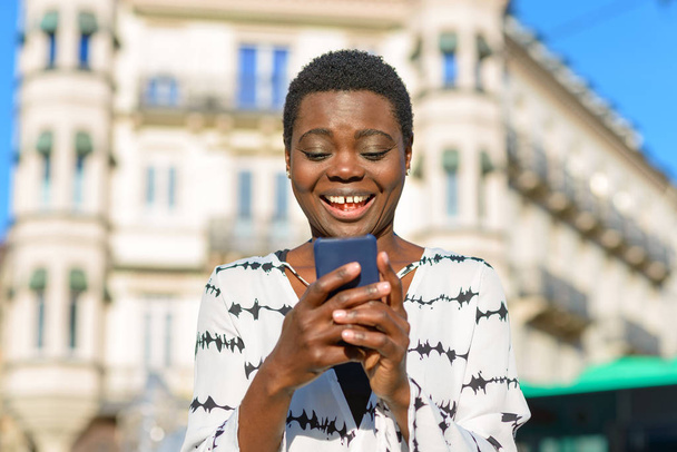 Felice elegante giovane donna africana ridendo del suo telefono cellulare mentre si trova in una strada della città in una vista a basso angolo contro un edificio storico
 - Foto, immagini