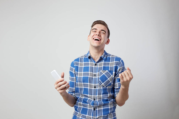 Ecstatic full of energy guy dressed in a plaid shirt keeps mobile phone in his hand a white background in the studio - Photo, image